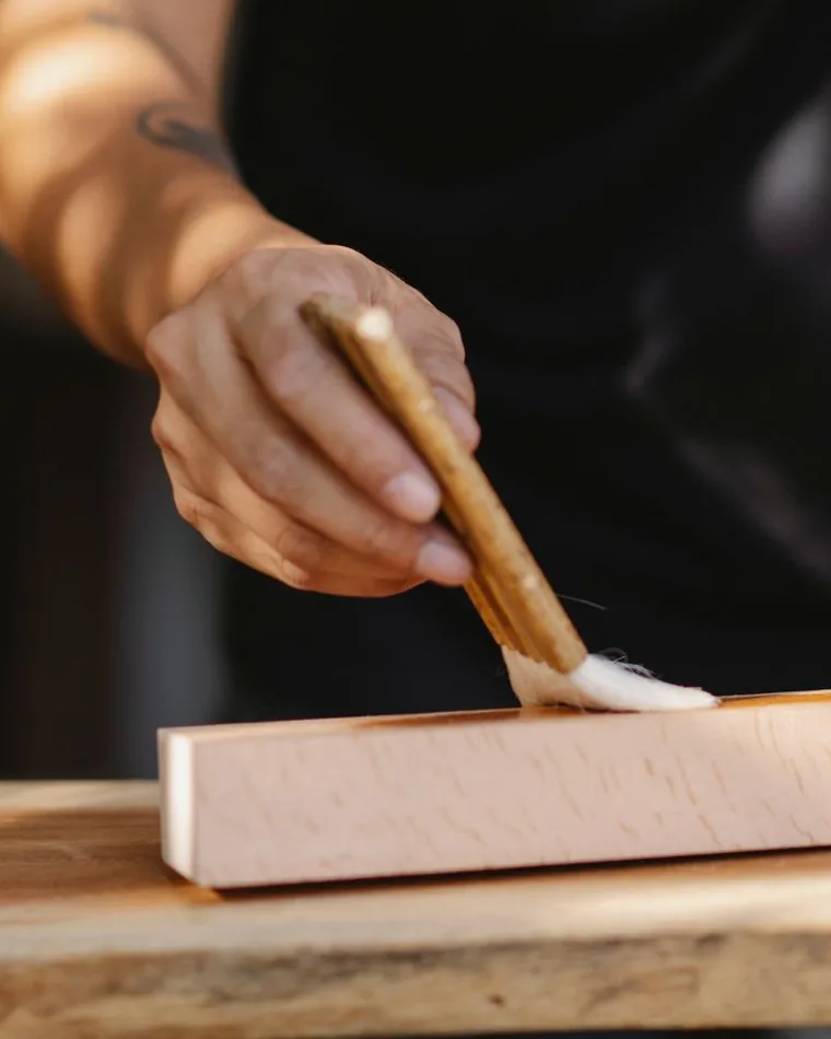 Aplicación de un acabado con brocha sobre un listón de madera clara apoyado en un banco de taller