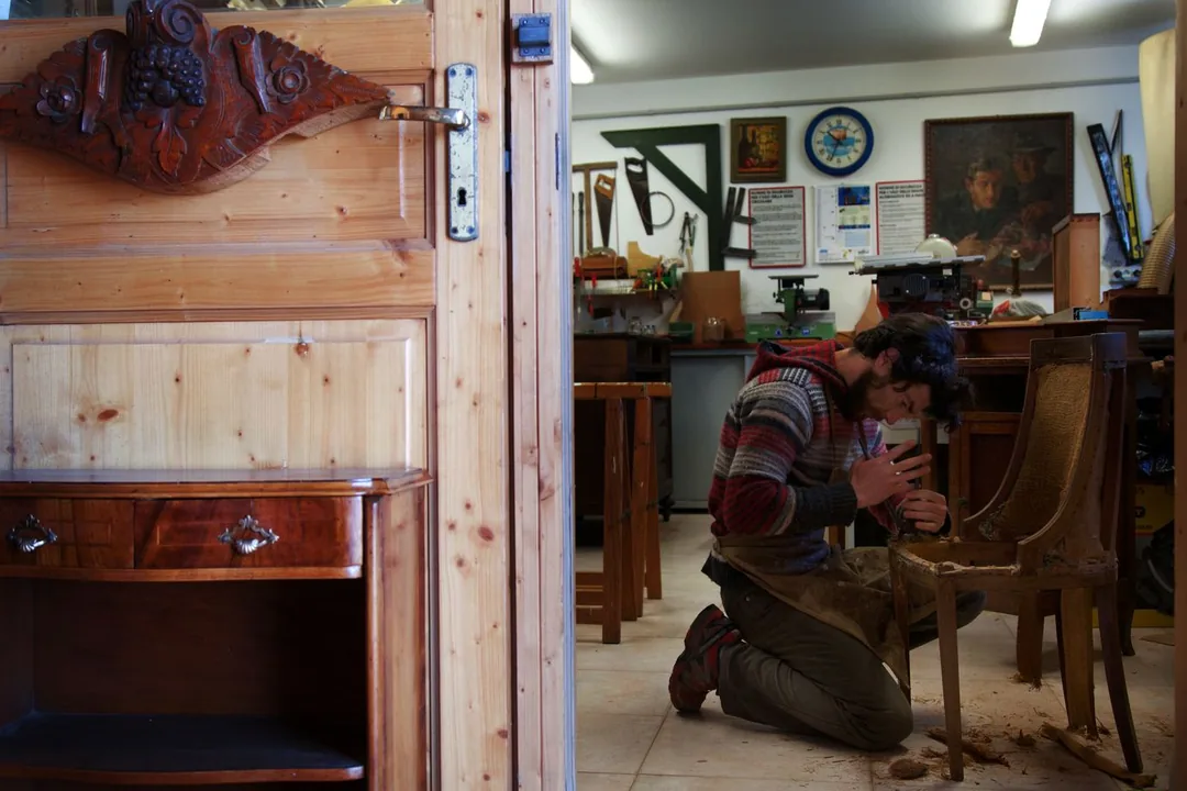 Interior de un taller de restauración con una silla antigua desmontada, virutas en el suelo y muebles de madera esperando reparación